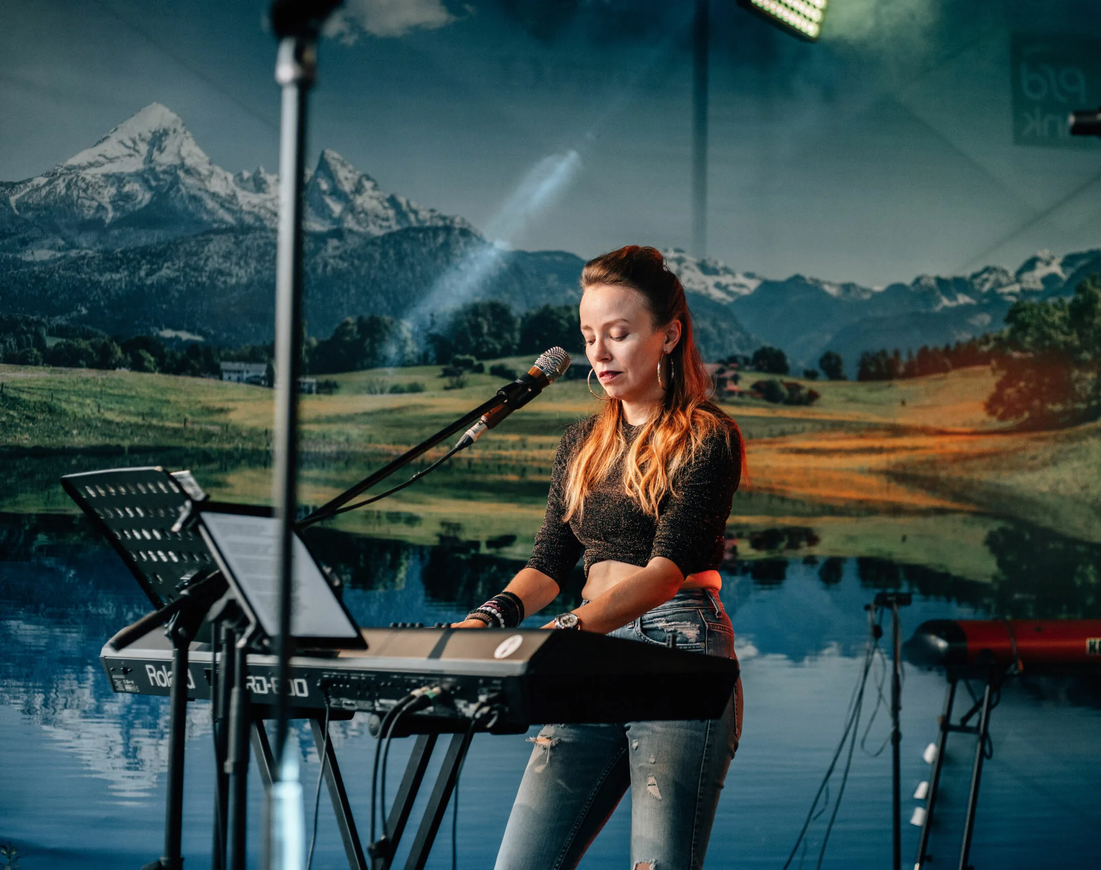 Miriam Geissler spielt bei einem Konzert auf der Bühne Keyboard, vor ihr ein Mikrofon, im Hintergrund ein Bergpanorama